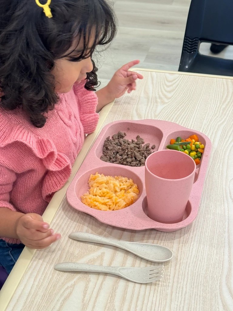A toddler at the table with a lunch tray of rice, beef and mixed vegetables